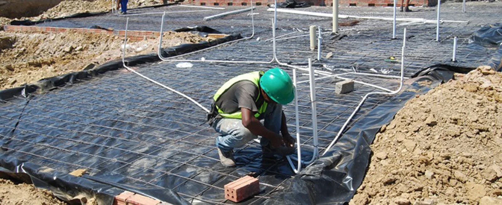 An installer kneeling on a foundation slab among rows of white conduit, before the screed is poured.